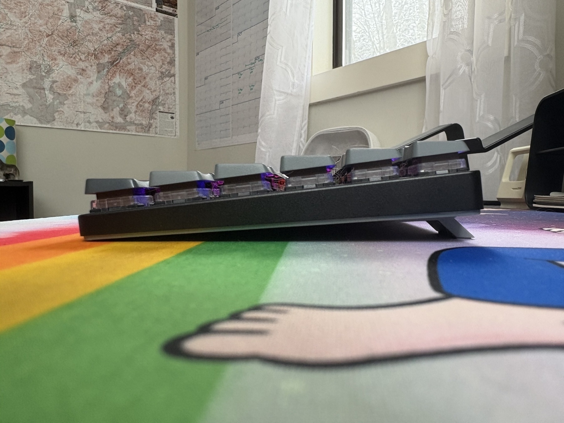 A mechanical keyboard rests at an angle, backlit with purple lights, on a colorful, cartoon-illustrated mat. In the background, maps hang on the walls near a window with sheer curtains.