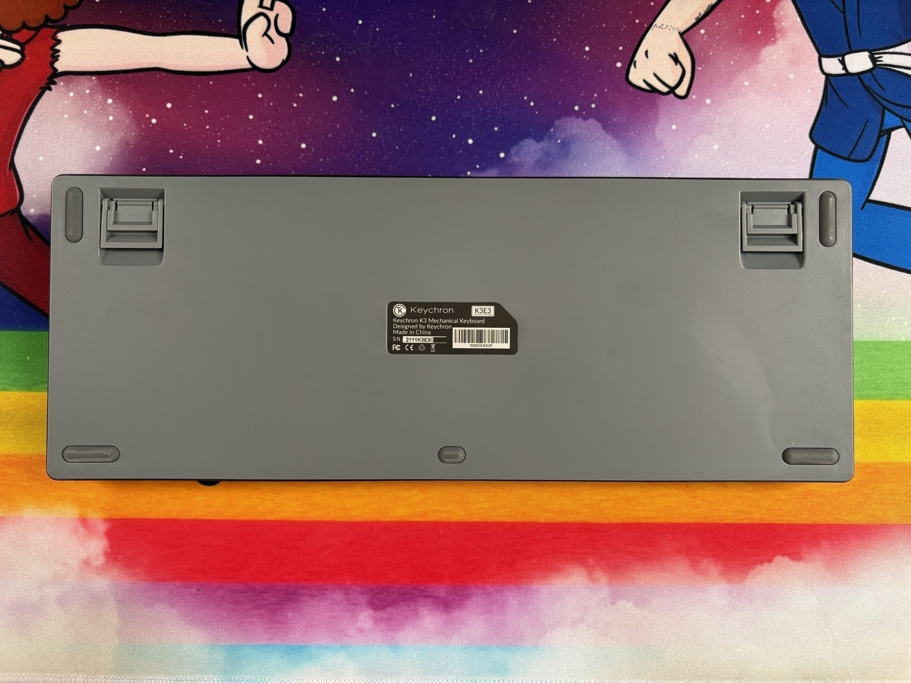 A gray keyboard rests upside down with adjustable feet extended, labeled “Keychron K3E3 Mechanical Keyboard.” It sits on a colorful mat featuring cartoon arms and rainbow stripes.