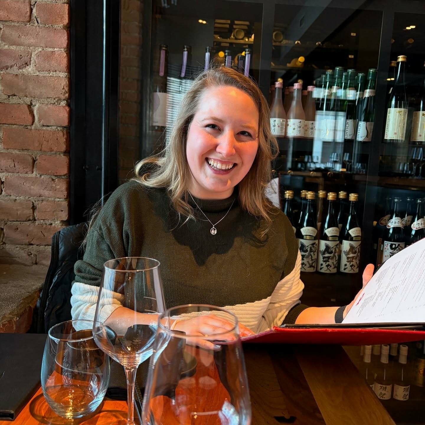 A woman smiling while holding a menu sits at a table with empty glasses in a restaurant. Behind her, shelves display numerous bottles with various labels.
