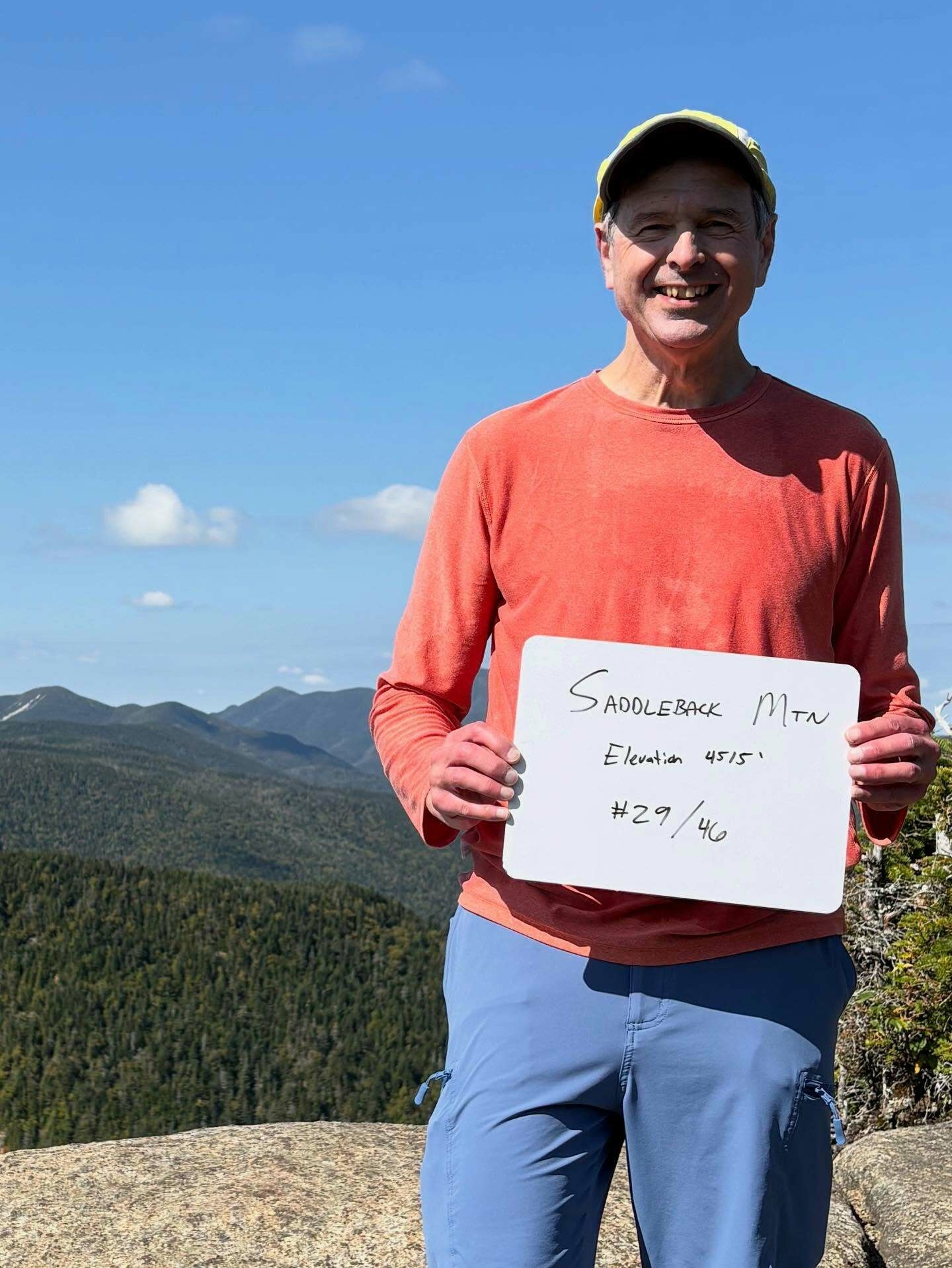 A person holding a white sign stands smiling on a rocky mountain summit with tree-covered hills in the distance under a clear blue sky.
Text on the sign: “Saddleback Mtn Elevation 4515' #29/46”.
