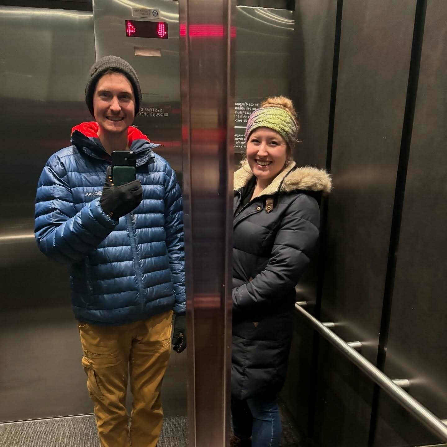 Two people wearing winter coats and hats standing inside an elevator looking at their reflection in the mirror with metallic walls and railings surrounding them.
