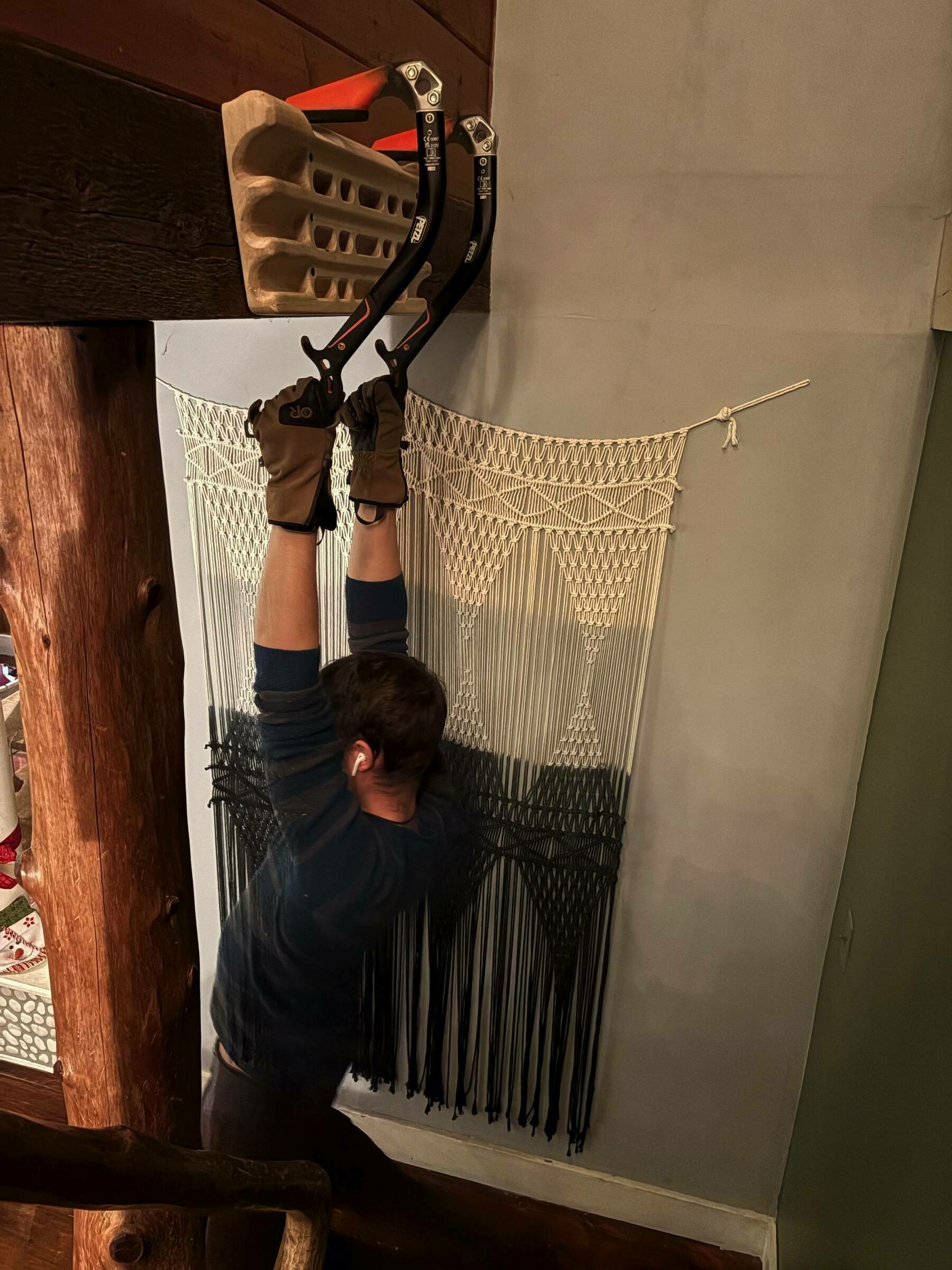 A person wearing gloves hangs from two ice tools attached to a climbing hold, set against an interior wall decorated with macramé art.