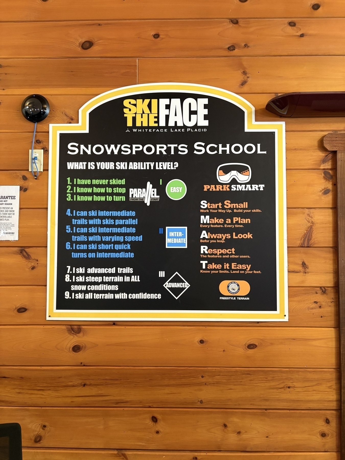 Signboard displays ski levels, safety tips at a snowsports school on a wooden wall. Text reads: Ski the Face, Whiteface Lake Placid, What is your ski ability level, and Park Smart guidelines.