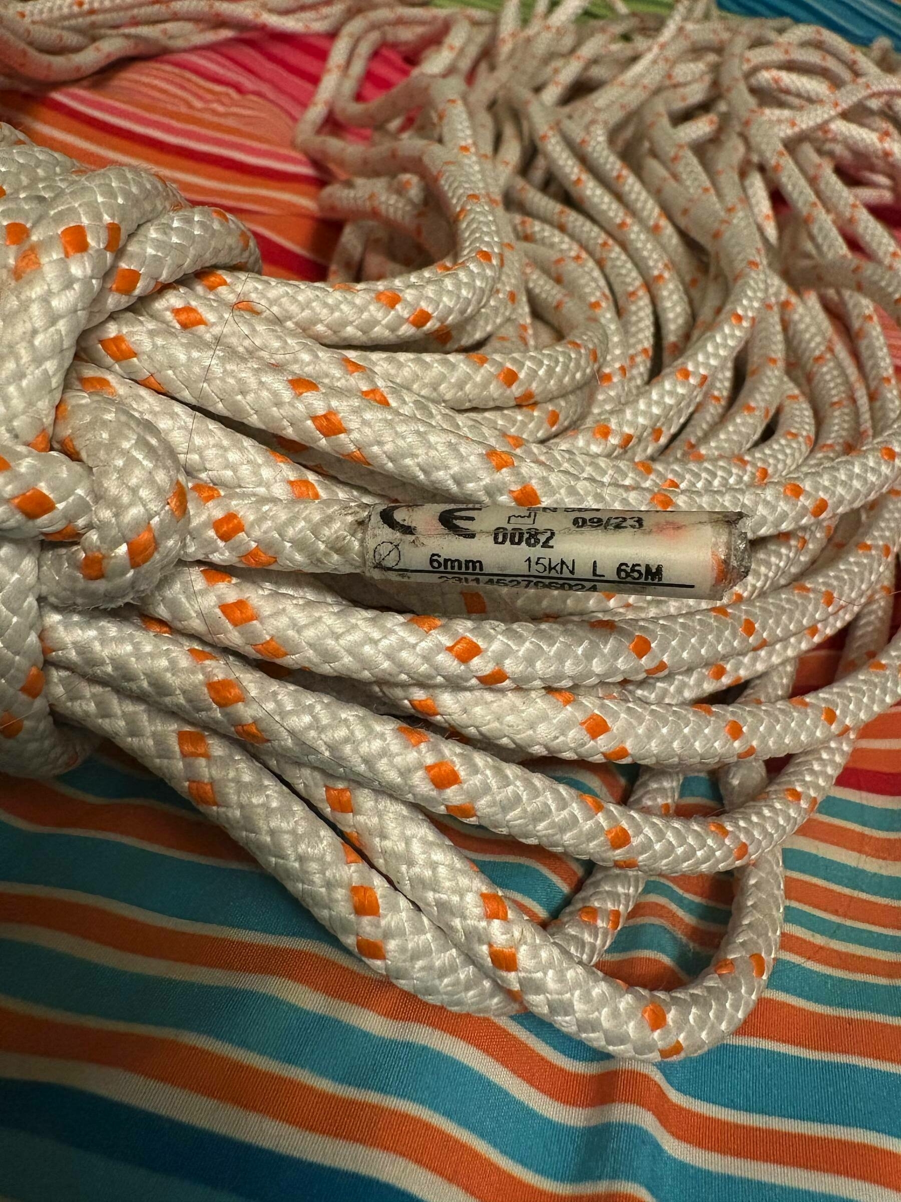 Coiled white rope with orange accents rests on a colorful striped fabric. Label indicates “6mm, 15kN, L 65m” with other markings, suggesting specifications and safety standards.