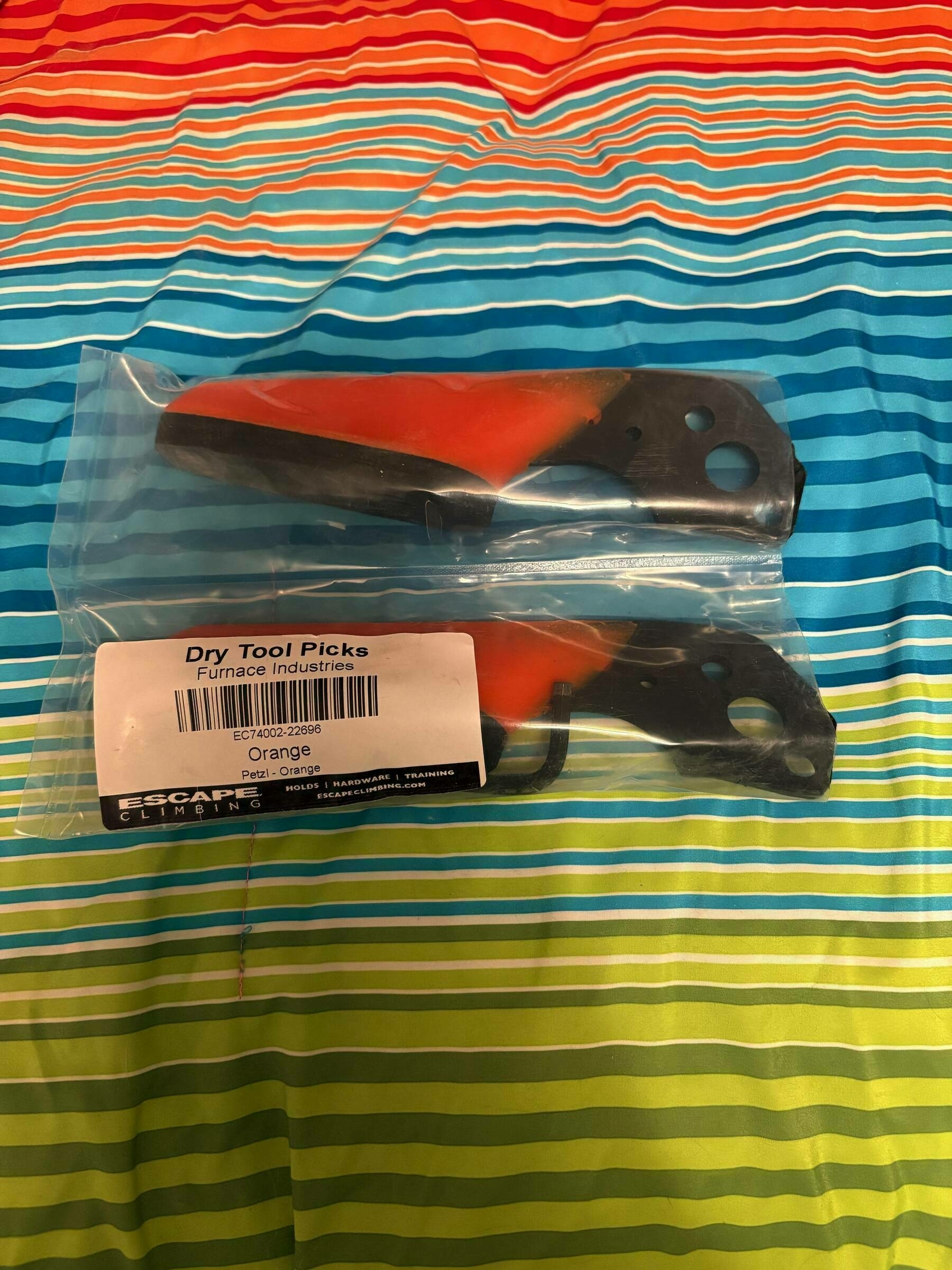 Two orange and black dry tool picks, encased in plastic packaging, rest on a vibrant, striped surface. Label reads: “Dry Tool Picks, Furnace Industries, Orange, Petzl - Orange, ESCAPE CLIMBING.”