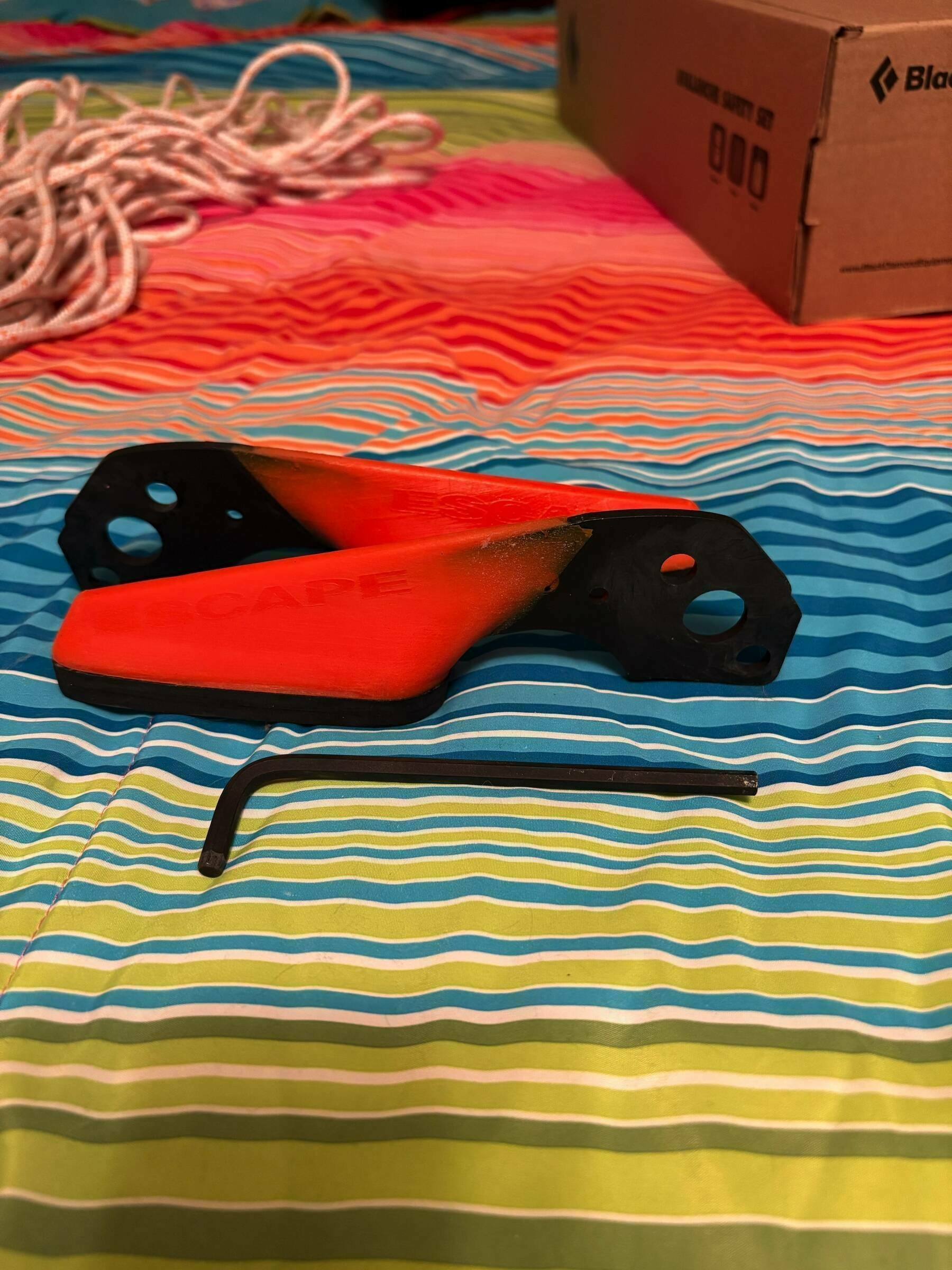 Orange climbing grips and an Allen wrench rest on a colorful, striped fabric surface. A coiled rope and a cardboard box are nearby. Text on the grip reads “ESCAPE.”