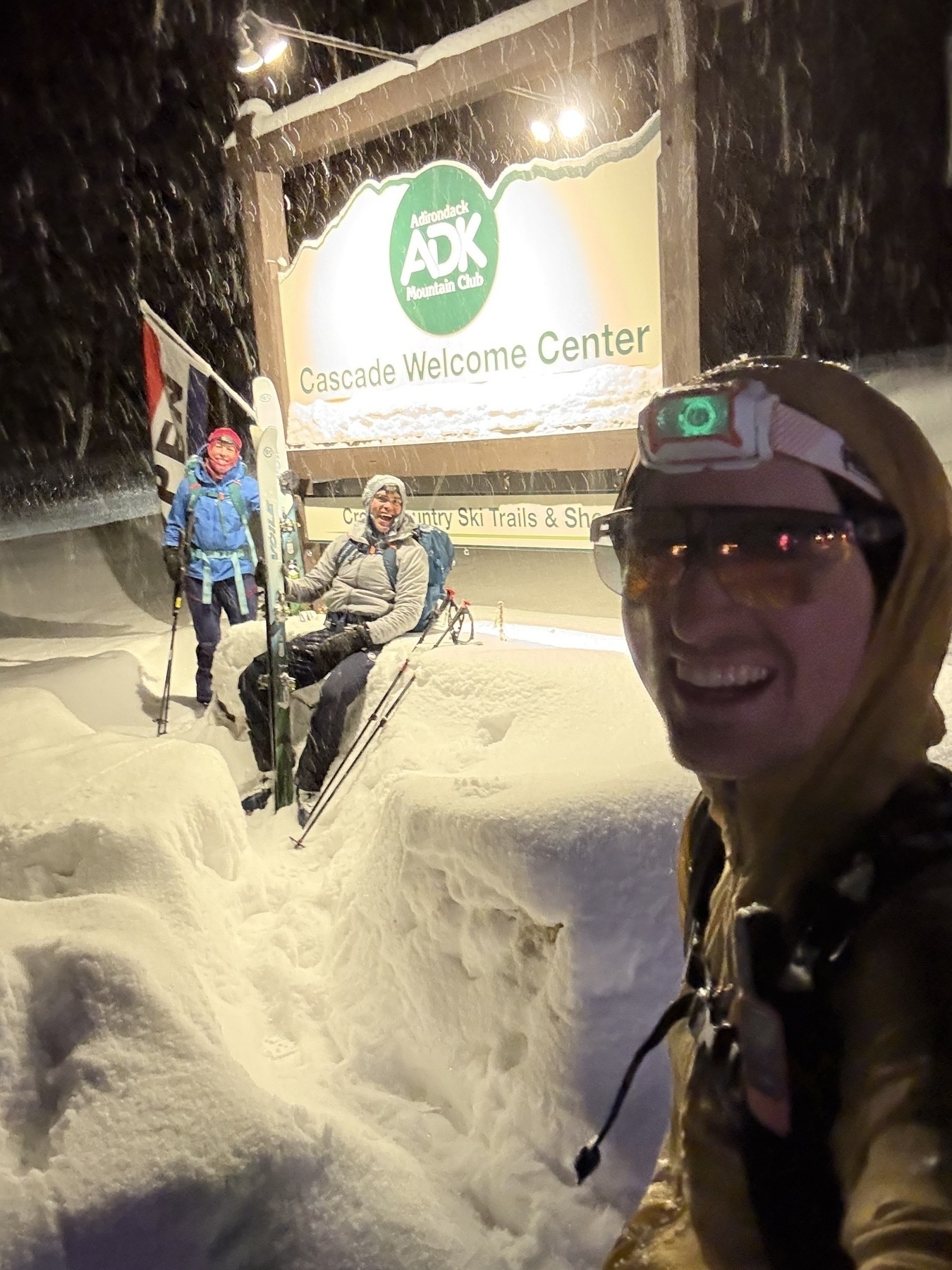 Three people pose happily in front of the Cascade Welcome Center sign, surrounded by deep snow, at night. The sign reads: Adirondack Mountain Club Cascade Welcome Center.