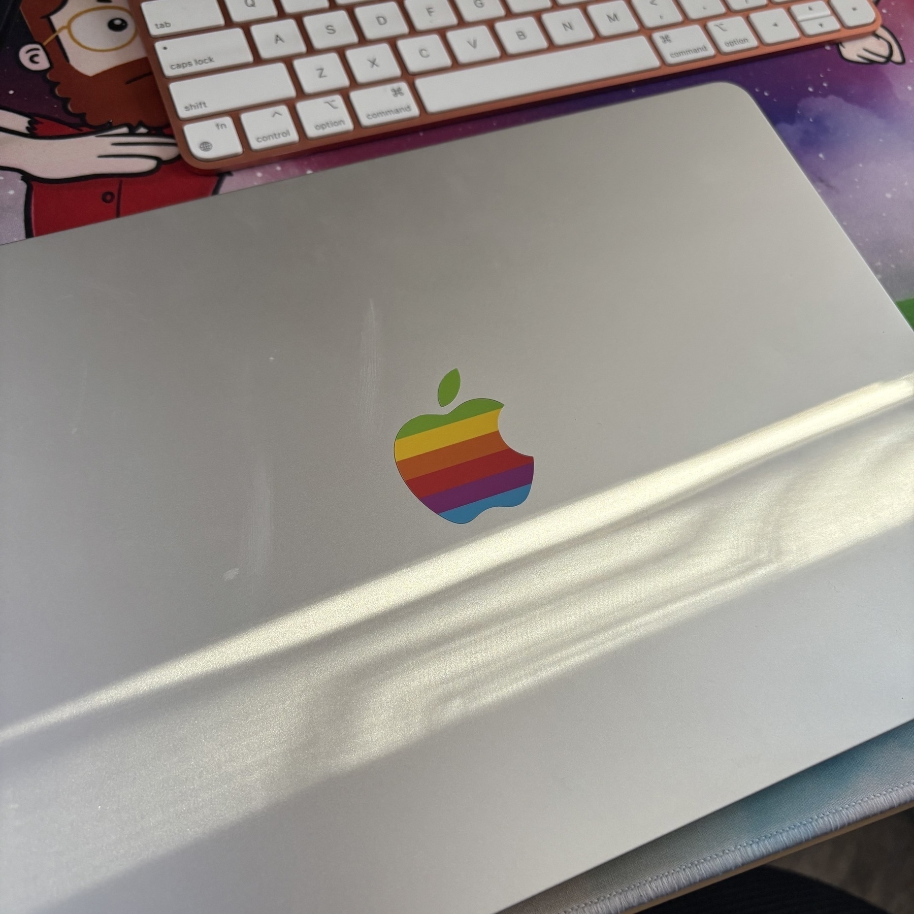 A close-up shot of a silver laptop with a retro rainbow-colored apple logo on it. Part of a keyboard is visible in the background along with a colorful mouse pad featuring a cartoon character design. Soft lighting casts a reflection on the laptop surface.