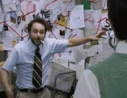 A man with disheveled hair is animatedly gesturing with both hands, holding a cigarette in his mouth. He stands in front of a chaotic wall covered with papers, notes, and red strings connecting various points. Another person is partially visible in the foreground, looking towards the man. The scene conveys a sense of urgency or conspiracy theory unraveling.