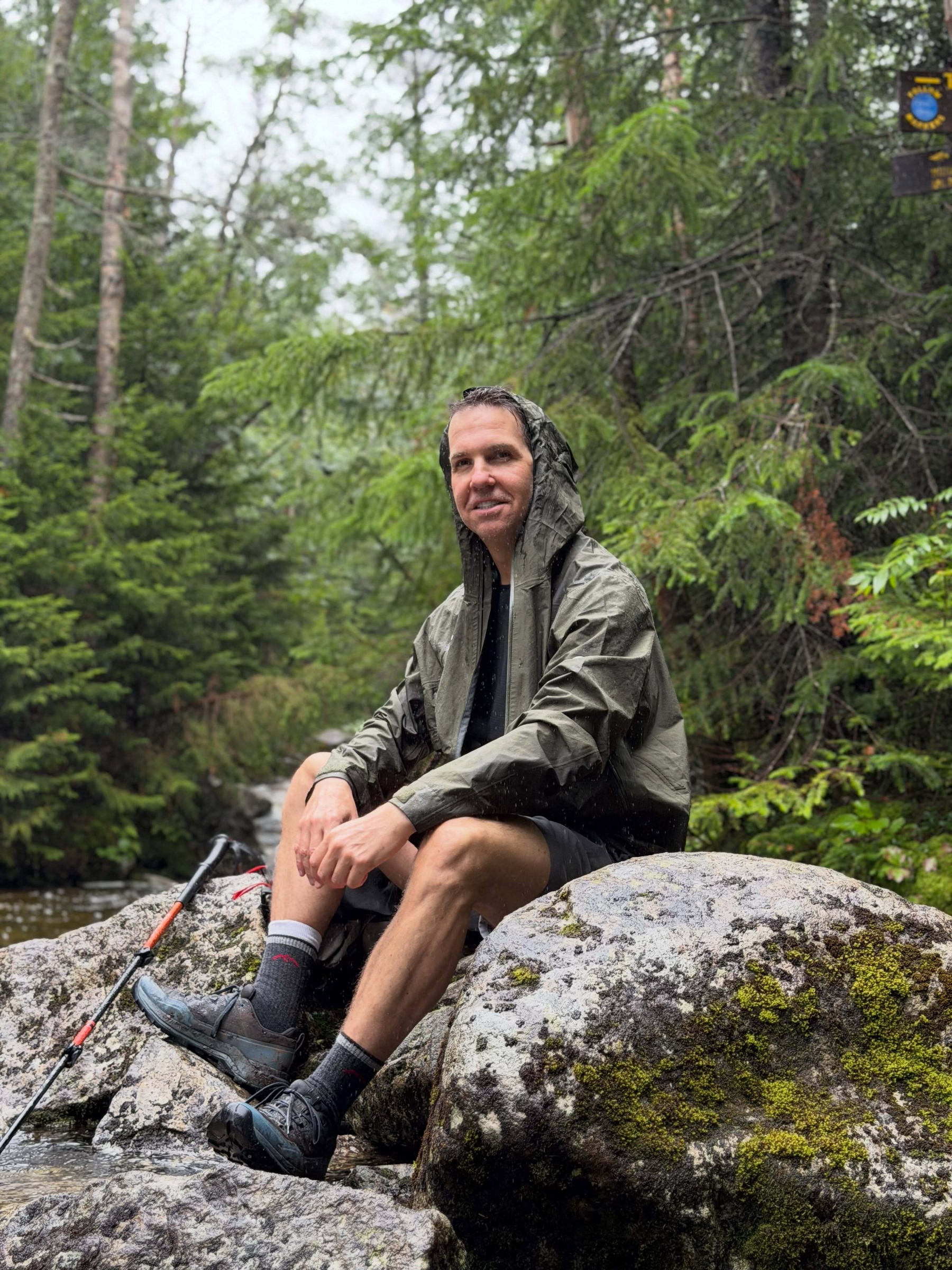 A person wearing a rain jacket sits on a mossy rock holding a hiking pole surrounded by a lush green forest near a stream.