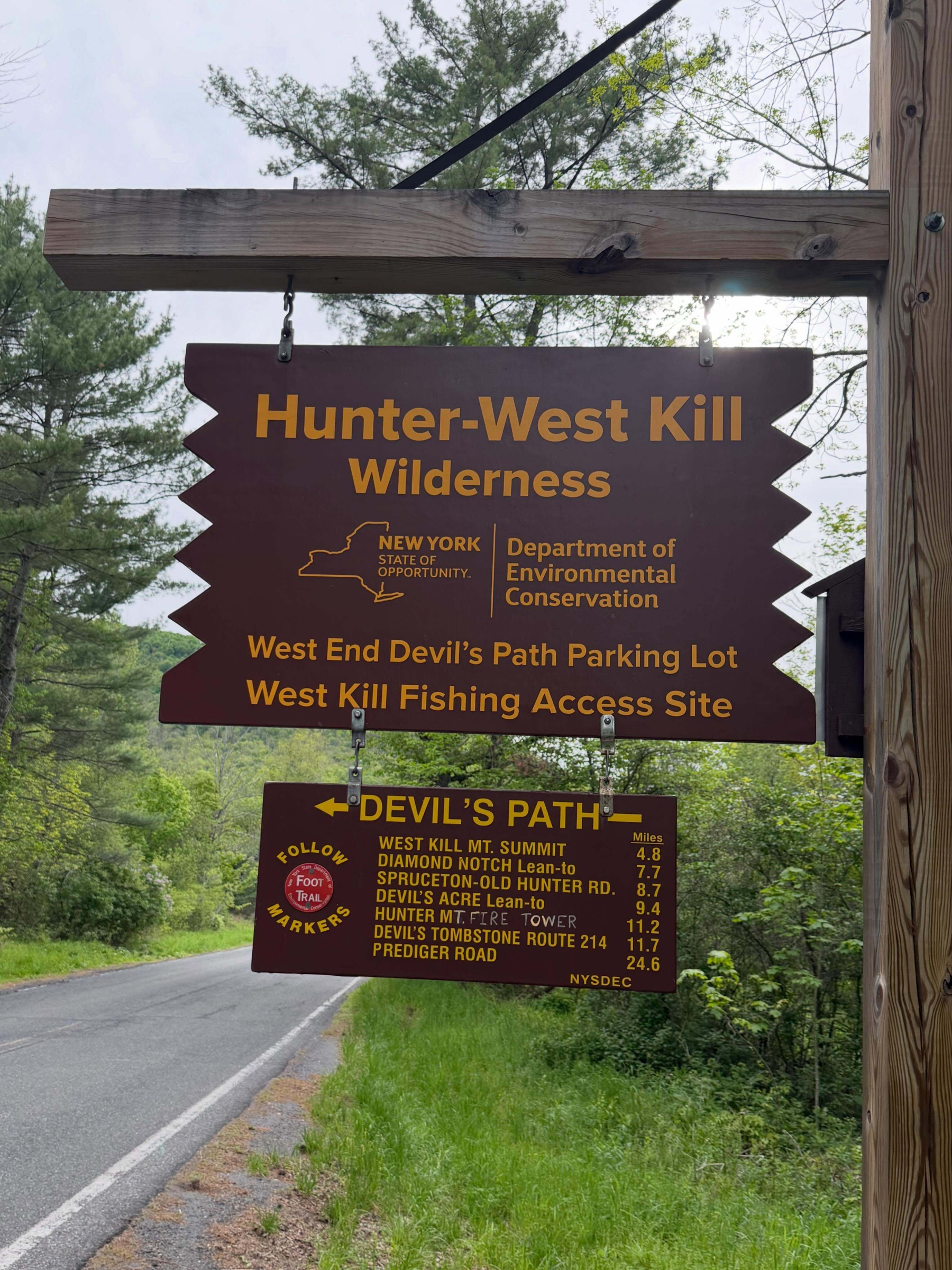 A wooden sign hanging beside a road indicates Hunter-West Kill Wilderness with trail directions. Text reads Hunter-West Kill Wilderness New York State Department of Environmental Conservation West End Devil&rsquo;s Path Parking Lot West Kill Fishing Access Site. Devil&rsquo;s Path Follow Foot Trail Markers West Kill Mt. Summit 4.8 Diamond Notch Lean-to 7.7 Spruceton-Old Hunter Rd. 8.7 Devil&rsquo;s Acre Lean-to 9.4 Hunter Mt. Fire Tower 11.2 Devil&rsquo;s Tombstone Route 214 11.7 Prediger Road 24.6 NYSDEC. Surrounded by trees.