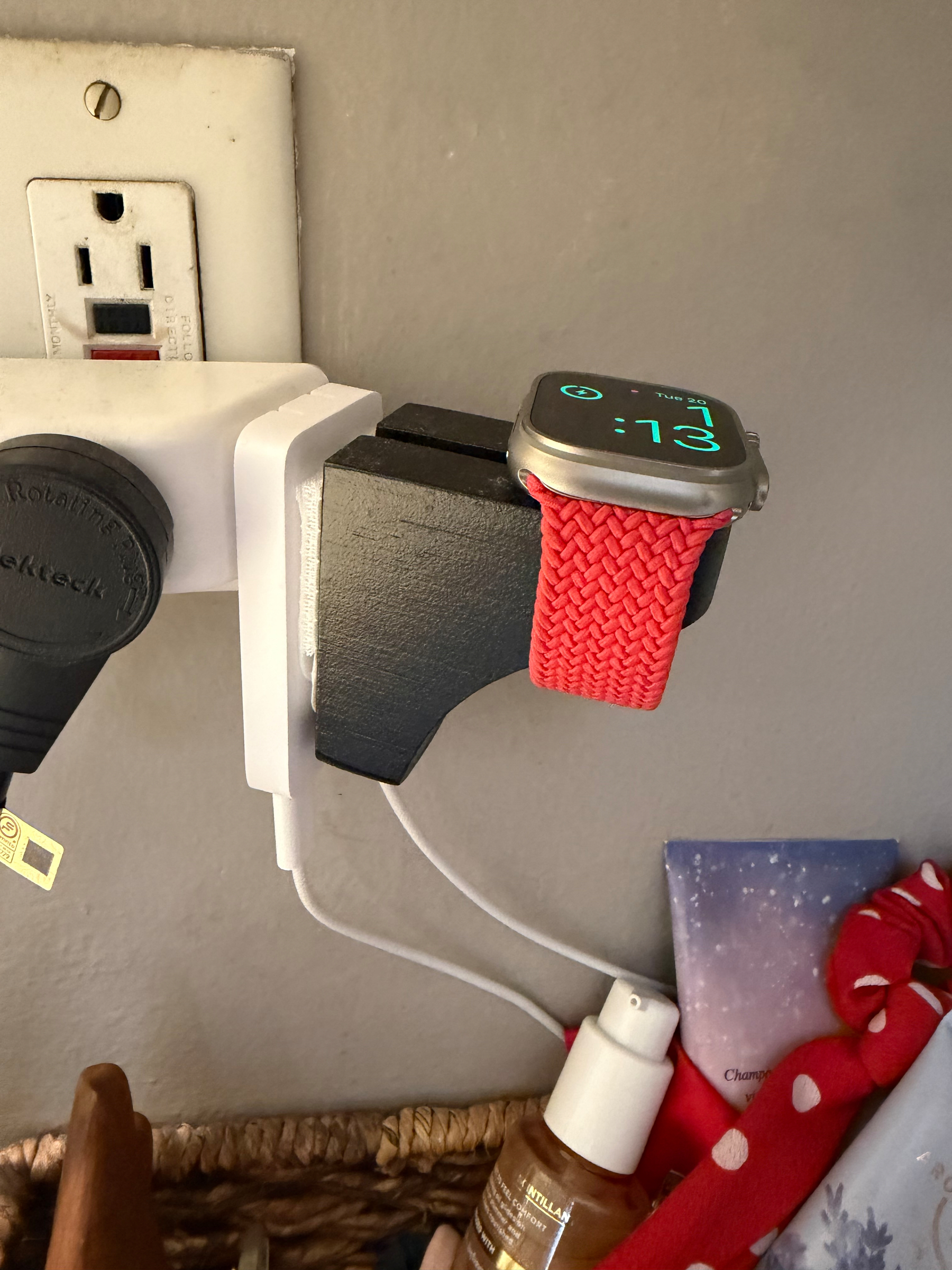 A smartwatch with a red woven band is displayed on a charging stand attached to an electrical outlet. The watch face shows the time as 7:13. There’s a USB cable plugged into the stand. A wicker basket below contains a spray bottle, a tube of skincare product, and a red item with white polka dots.