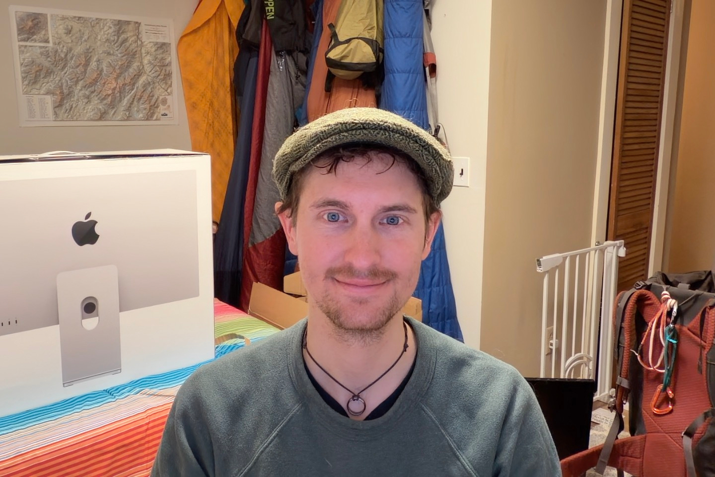 A person wearing a flat cap and a necklace is smiling at the camera. They are seated in a room with an iMac box and colorful camping gear visible in the background. A wall map is partially seen on the left, and a backpack with carabiners is on the right.